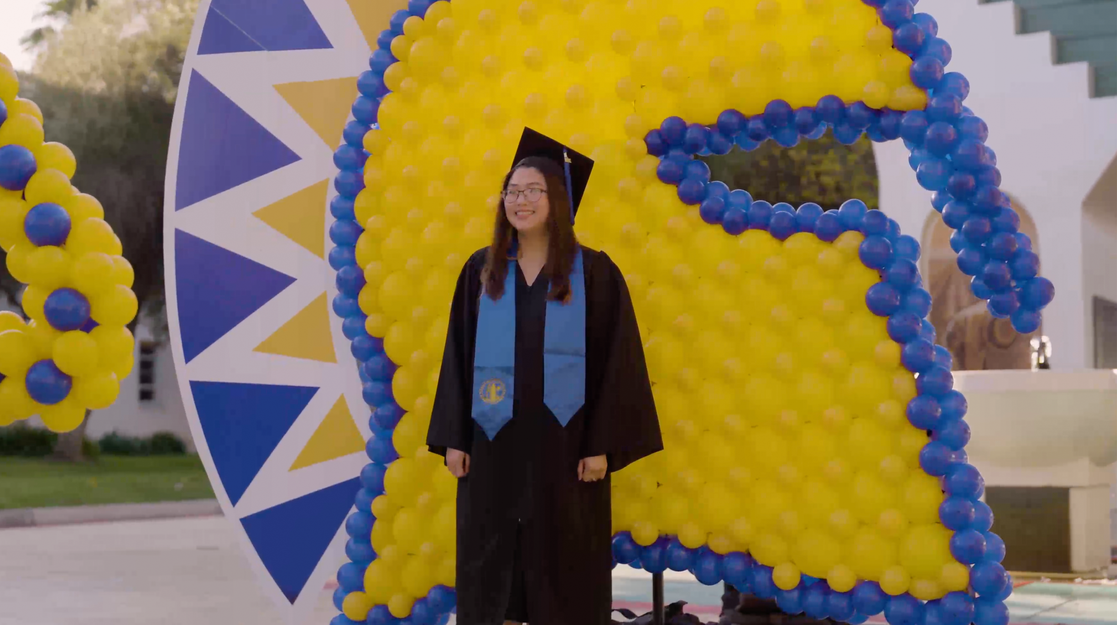 Still of a graduate gettting their photo taken at a balloon art spartan backdrop